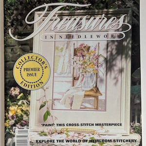 Op de afbeelding: Een tijdschriftomslag getiteld "Treasures in Needlework" toont een ingelijst kruissteekontwerp van bloemen in een kan op een stoel. De cover bevat de tekst "Collector's Premier Issue Edition" en "Explore the World of Heirloom Stitchery."