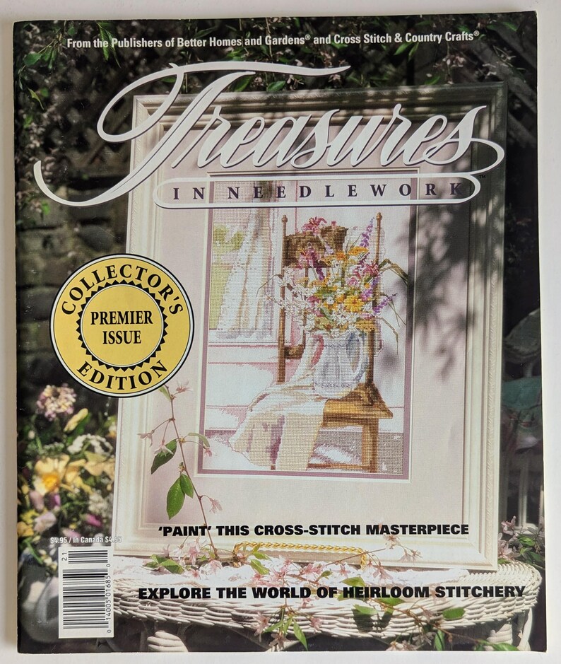 Op de afbeelding: Een tijdschriftomslag getiteld "Treasures in Needlework" toont een ingelijst kruissteekontwerp van bloemen in een kan op een stoel. De cover bevat de tekst "Collector's Premier Issue Edition" en "Explore the World of Heirloom Stitchery."