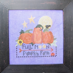 Op de afbeelding: Kruissteekkunst in een donkerhouten lijst. Het ontwerp toont oranje pompoenen, een volle maan en sterren op een lavendelkleurige achtergrond. De tekst luidt "Full Moon Pumpkin Farm."