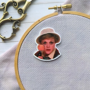 Puede incluir: Una pegatina blanca con una imagen de dibujos animados de una mujer con un sombrero y una lupa. La mujer tiene el pelo rubio y mira a la cámara con una expresión seria. La pegatina está sobre un fondo de tela gris claro.