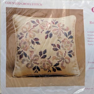 Puede incluir: Un kit de punto de cruz contado con un cojín beige con un diseño floral. El cojín tiene un patrón cosido de rosas y hojas en tonos de rosa, marrón y azul. El tamaño final es de 30,48 cm x 30,48 cm. El kit incluye tela Aida de 14 hilos.