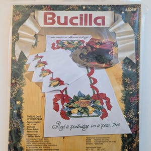 Puede incluir: Un kit de camino de mesa para bordar en punto de cruz Bucilla titulado Twelve Days of Christmas, con un diseño de perdiz en un peral. El paquete incluye tela impresa, hilo de bordar 100% algodón, una aguja e instrucciones para un camino de 35,5 por 111,7 cm.