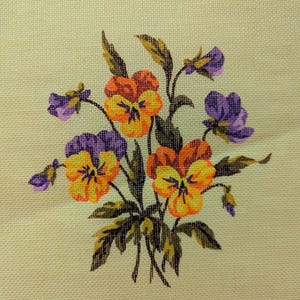 May include: A cross-stitch embroidery piece on cream-coloured fabric featuring a bouquet of pansies. The flowers display vibrant shades of orange, yellow, and purple, accompanied by dark green leaves and stems, all rendered in detailed needlework.