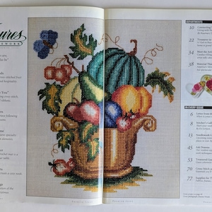 Op de afbeelding: Een kruissteekontwerp van een overvloedshoorn vol fruit en groenten, waaronder een watermeloen, pompoenen en druiven. De afbeelding komt uit een tijdschrift getiteld "Treasures in Needlework."