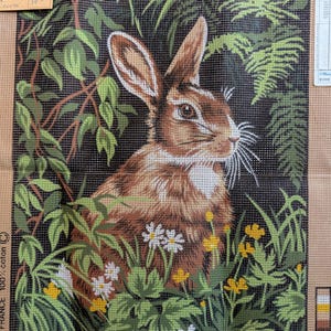 May include: A needlepoint canvas featuring a brown rabbit surrounded by green foliage, ferns, and small white and yellow flowers. The design is printed on a tan grid background with a colour key on the side and the text Creations Margot de Paris.