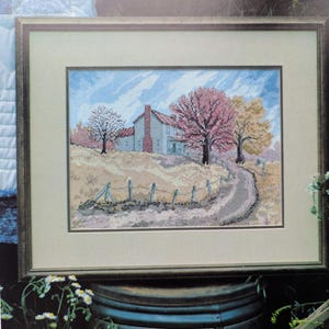 Könnte beinhalten: Gerahmte Kreuzstich-Landschaft mit einem Haus mit rotem Backsteinkamin, einer gewundenen Straße und Bäumen mit rosa und gelbem Laub. Das Kunstwerk ist vor blauem Himmel in einem braunen Holzrahmen gerahmt.