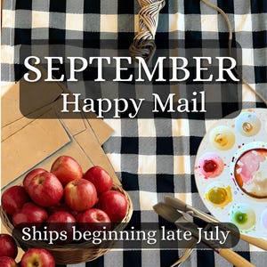 May include: A woven basket filled with red apples, a white paint palette with colorful paint wells, and two paintbrushes arranged on a black and white buffalo check fabric. The text reads SEPTEMBER Happy Mail and Ships beginning late July.