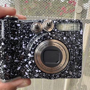 以下が含まれることがあります： 白のペイントスプラッター模様が施された黒いデジタルカメラ。Canon Zoom Lens 4x 5.8-23.2mm 1:2.6-5.5レンズ、前面上部に取り付けられた2つの銀色の金属製トグルスイッチ、および長方形のフラッシュユニットを備えています。