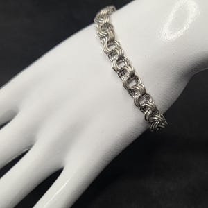 May include: A silver-colored bracelet with a chain-link design. The bracelet is displayed on a white mannequin hand against a black background. The links are closely connected, creating a textured appearance. The bracelet is a jewelry item.
