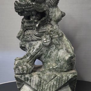 May include: A detailed sculpture of a mythical creature, possibly a guardian lion, carved from green and gray stone. The statue is seated on a geometric base, showcasing intricate details and textures.