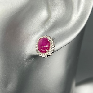 Op de afbeelding: Enkele oorbel met een ovale robijn edelsteen, omringd door een halo van kleine, heldere, sprankelende stenen. De zetting is zilverkleurig. De oorbel wordt getoond op een mannequin oor.