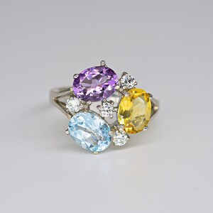 Könnte beinhalten: Ein silberner Ring mit einer Anordnung farbiger Edelsteine. Der Ring zeigt einen oval geschliffenen lila Amethysten, einen gelben Citrin und einen hellblauen Aquamarin, akzentuiert mit kleinen, runden, klaren Steinen. Die Edelsteine sind in einem Cluster-Design angeordnet.