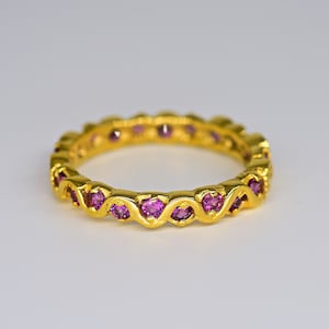 Puede incluir: Un anillo dorado con un patrón repetitivo de piedras preciosas rosas. Las piedras están engastadas en un diseño ondulado alrededor de la banda. El anillo se muestra sobre un fondo blanco.