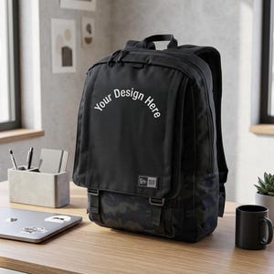 Könnte beinhalten: Schwarzer Rucksack mit Klappe und Camouflagemuster am Boden. Der Text "Your Design Here" ist auf die Klappe gedruckt. Der Rucksack hat verstellbare Träger und Schnallen. Eine schwarze Tasse und ein Laptop stehen auf dem Schreibtisch.