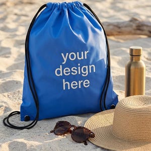 May include: A blue drawstring bag with the text "your design here" in white, a pair of sunglasses, a straw hat, and a gold-coloured water bottle on a sandy beach. The bag has black drawstrings.