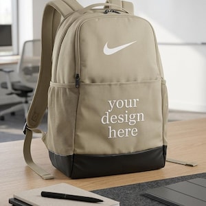 May include: Tan backpack with a white Nike logo and the text "your design here." The backpack has black accents, mesh side pockets, and a top handle. A stack of notebooks and a pen are in the foreground.