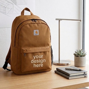 Peut inclure: Un sac à dos marron avec une poche avant et le texte "your design here" en blanc. Le sac à dos a une poignée supérieure, des bretelles réglables et un petit écusson logo. Une poche en filet est visible sur le côté.