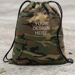 Op de afbeelding: Camouflage trekkoordtas met een voorvak en zwarte touwriemen. De tas heeft de tekst "YOUR DESIGN HERE" in het wit. De tas is gemaakt van een camouflagepatroon in tinten groen, bruin en beige.