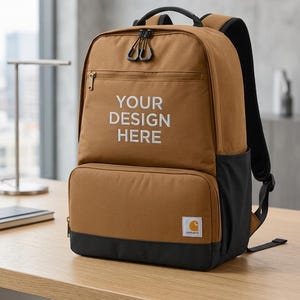 May include: Brown backpack with black accents and the text "YOUR DESIGN HERE" in white. Features a zippered front pocket, a top handle, and padded shoulder straps. The Carhartt logo is visible on the lower right corner.