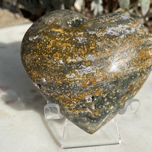 May include: A heart-shaped stone sculpture with a mottled pattern of gray, gold, and white. The polished surface of the stone is displayed on a clear acrylic stand, showcasing its natural beauty. The sculpture is a unique decorative item.