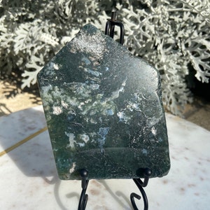 May include: A green moss agate slab with white and gray inclusions, displayed on a black metal stand.