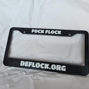 Puede incluir: Marco de matrícula negro con el texto "FOCK FLOCK" en la parte superior y "DEFLOCK.ORG" en la parte inferior en letras mayúsculas blancas. El marco tiene forma rectangular con esquinas redondeadas y dos orificios de montaje.