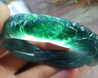 May include: A translucent green jade bracelet with a floral design. The bracelet is carved with detailed flower patterns and has a smooth, polished finish. The jade is a deep green color, with some areas appearing lighter due to the light passing through.