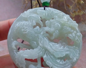 May include: A round, pale green jade pendant with a carved phoenix and dragon design. The pendant is suspended from a dark cord with decorative beads. The intricate carving showcases the mythical creatures in a detailed, flowing style.