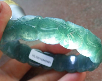 May include: A green jade bangle bracelet with a carved floral design. The bracelet is made of a translucent jade stone and has a polished finish. The bracelet is approximately 6 inches in diameter.