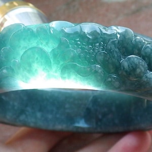Puede incluir: Un brazalete de jadeíta tallado con un diseño floral detallado. El brazalete es de color verde claro con motas blancas.
