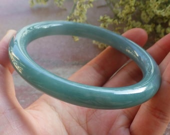 May include: A jade green bangle bracelet with a smooth, polished finish. The light reflects off the surface, showing the colour variations. This classic piece of jewellery is suitable for everyday wear or special events. The bracelet's diameter is approximately 6.5 cm.