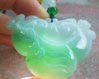May include: A jade pendant in shades of green, with a smooth, polished surface. The pendant has a unique, organic shape, possibly depicting a stylized figure or form. The jade is translucent, allowing light to pass through, and is held by a string.