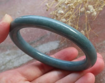 May include: A teal-green jade bangle bracelet. The smooth, rounded band of the bracelet displays a muted teal-green colour. The polished surface reflects light, showing subtle colour variations. The jewellery is held in a hand.