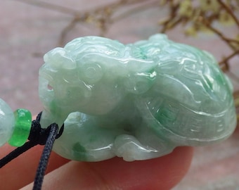 May include: A carved jade pendant in shades of green and white, depicting a mythical creature. The pendant is intricately detailed with a smooth, polished finish. A black cord and jade beads are attached.