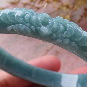 May include: A jadeite jade bangle bracelet in a soft teal color. The bracelet features carved floral and leaf designs along the top. The jade is translucent, with visible veining. A beautiful piece of jewelry.
