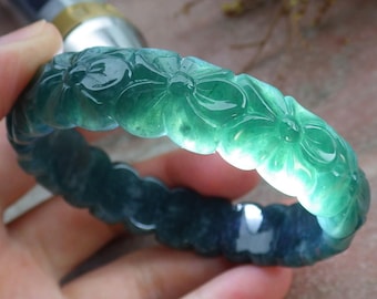 May include: A jade bracelet with a floral design. The bracelet is a gradient of green and dark teal. The flowers are carved into the surface of the bracelet.