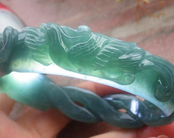 May include: A translucent green jade bangle bracelet featuring intricate relief carvings of a phoenix with detailed wing feathers and swirling cloud motifs. The stone displays varying shades of deep and light green with a smooth, polished surface.