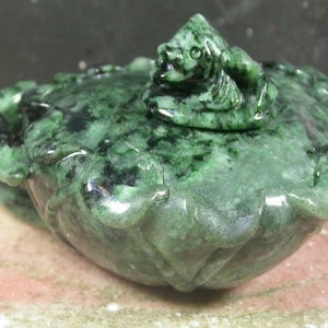 Puede incluir: Una tetera de jade verde con un remate de pez. La tetera está tallada en forma de flor de loto.