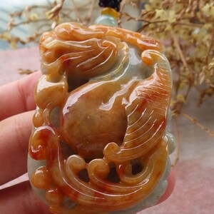 Può includere: Un ciondolo in giada scolpito a forma di creatura mitologica con dettagli intricati. Il ciondolo è di colore verde chiaro con accenti rossi e arancioni.