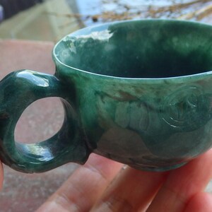 Puede incluir: Taza de té verde jade hecha a mano con un diseño de asa único. La taza está tallada en una sola pieza de jade, mostrando las variaciones naturales de color y textura. El asa tiene una forma curva distintiva. La taza está sostenida en una mano.
