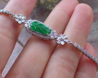 Op de afbeelding: Een zilveren armband met een centrale ovale groene jade steen, omringd door kleine heldere edelstenen. De armband heeft een delicaat ontwerp met bloemaccenten en een ketting van kleine, heldere stenen.