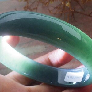 May include: A smooth, polished, circular jade bangle bracelet featuring a translucent deep green color with lighter emerald green variations. The bangle has a rounded profile and a glossy finish, with a small white rectangular sticker attached.