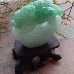 May include: A jade carving of two green dragons on a dark wooden stand. The dragons are intertwined and have a light green color.