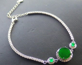 Op de afbeelding: Een zilveren armband met een grote, ronde, levendige groene edelsteen in het midden, geflankeerd door twee kleinere groene edelstenen. De armbandband is versierd met kleine, sprankelende heldere stenen en heeft een verstelbare kettingsluiting.