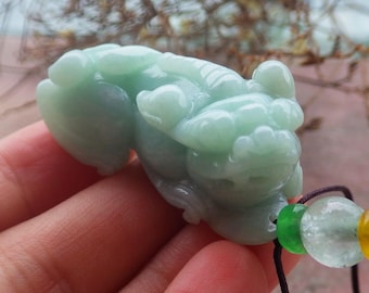 May include: A light green jade carving of a mythical creature, possibly a Pixiu, with intricate details. The carving is attached to a string with green, clear, and yellow beads. The jade has a smooth, polished finish.