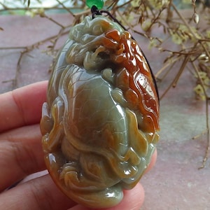 May include: A jade pendant with a carved design featuring a mythical creature. The pendant is primarily green and brown with orange accents. It hangs from a black cord with a green bead.