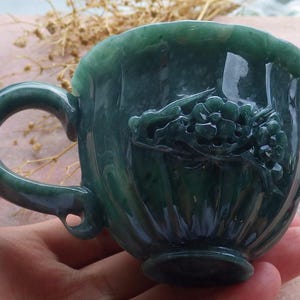 May include: A dark green jade teacup with a handle and a carved floral design. The cup has a glossy finish and a scalloped rim. The handle is rounded and attached to the side of the cup.
