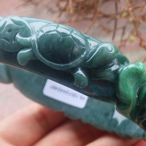 May include: A jade bangle bracelet in shades of green, featuring carved turtle designs. The bracelet has a smooth, polished finish and is held by a hand. The design includes detailed turtle figures and floral accents.