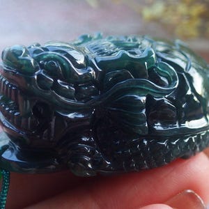 以下が含まれることがあります： 濃い緑色の翡翠で彫られた、龍亀の形をしたペンダント。ペンダントは非常に精巧に作られており、鋭い歯を持つ龍の頭と、複雑な彫刻が施された亀の甲羅が特徴です。