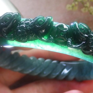 Könnte beinhalten: Ein grünes Jade-Armband mit aufwendigen Schnitzereien von Fischen und floralen Mustern. Das Armband ist poliert und hat eine glatte, glänzende Oberfläche.